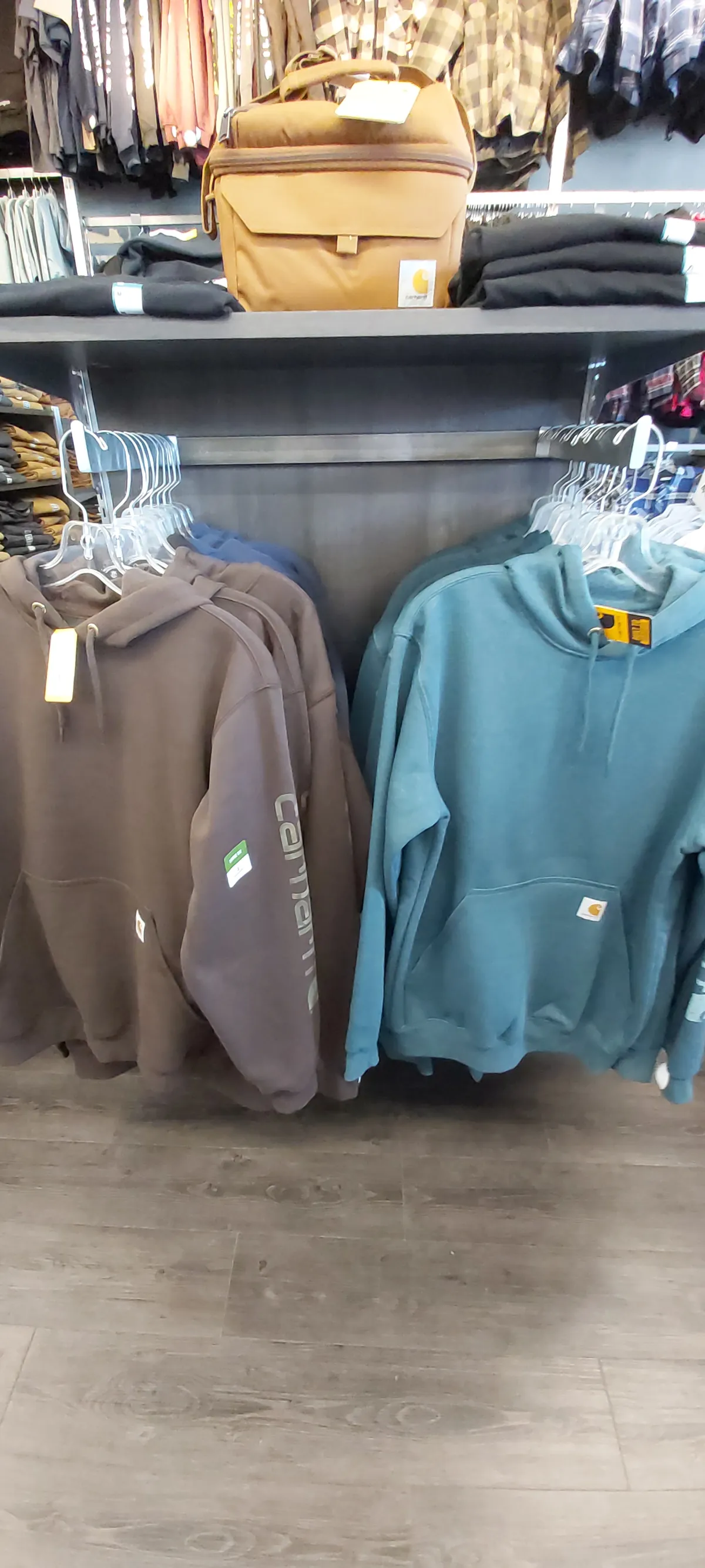 Carhartt hooded sweatshirts on a rack in teal and brown, bags on the shelf above